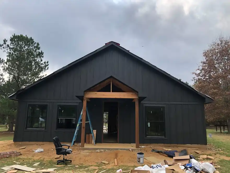 Black board-and-batten barndominium mid-job, ladder and drop cloths at the porch